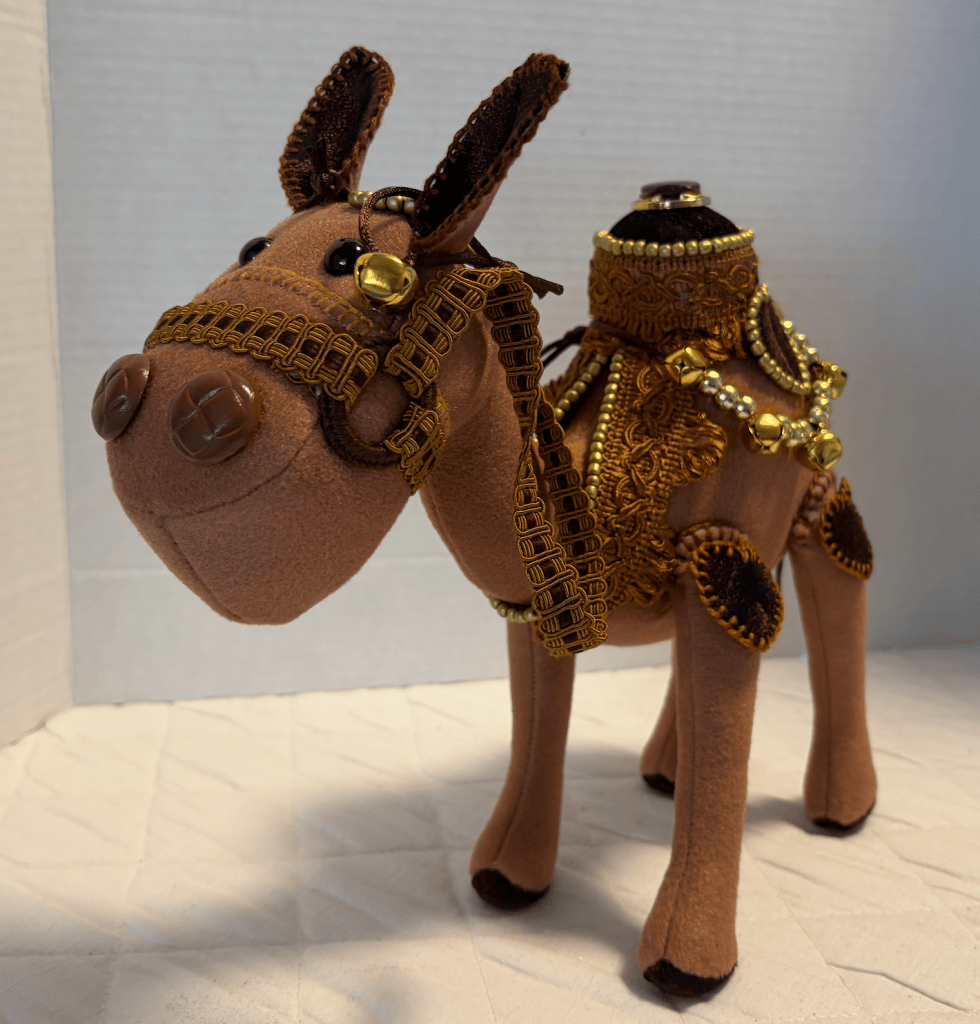 A handcrafted plush camel with a decorative saddle and golden accents, standing on a light-colored surface.