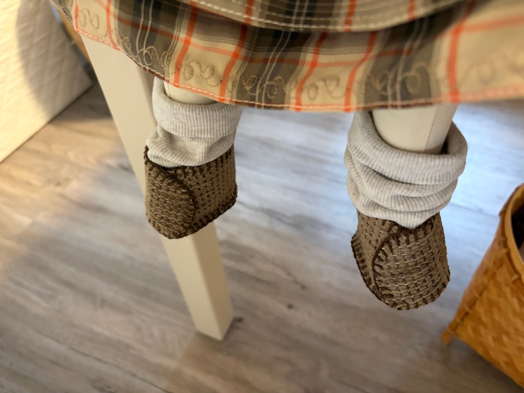 Close-up view of small feet in woven brown slippers, with light gray leggings. The feet dangle under a table, and part of a patterned blanket is visible in the background.