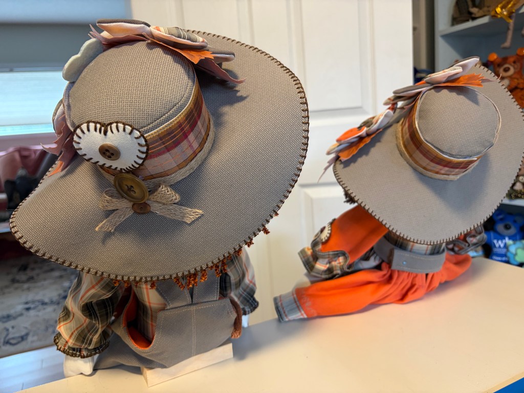 Two decorative figurines wearing large, stylish hats adorned with flowers and buttons, showcasing colorful plaid outfits, positioned on a shelf against a light background.