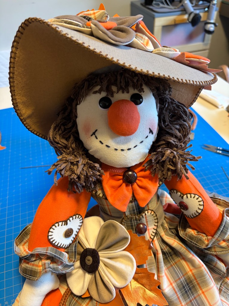 A close-up of a decorative scarecrow doll with a brown hat and a colorful dress. The doll features a smiling face, orange nose, and curly yarn hair, adorned with fabric flowers and buttons.