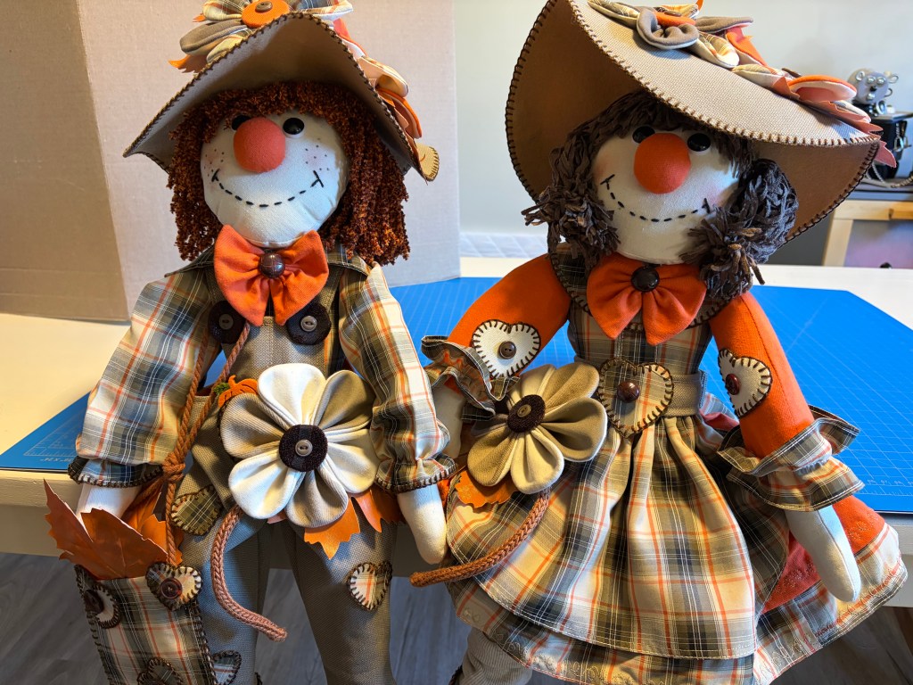 Two decorative scarecrow dolls with happy faces, dressed in plaid outfits adorned with flowers and bows, standing side by side.