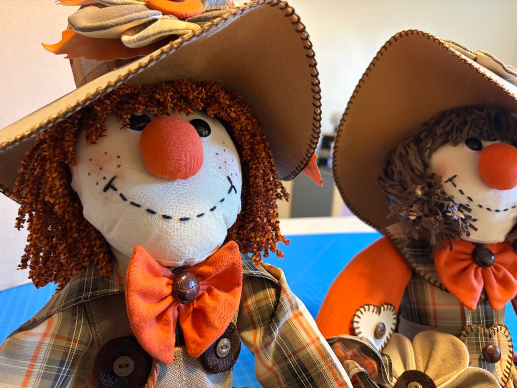 Two festive scarecrow dolls with orange bows, hats, and cheerful faces, sitting on a table against a colorful background.